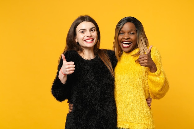 Foto dois amigos de mulheres jovens europeus e afro-americanos em roupas amarelas pretas em pé posando isolado no fundo da parede laranja brilhante, retrato de estúdio. conceito de estilo de vida de pessoas. mock up espaço de cópia.