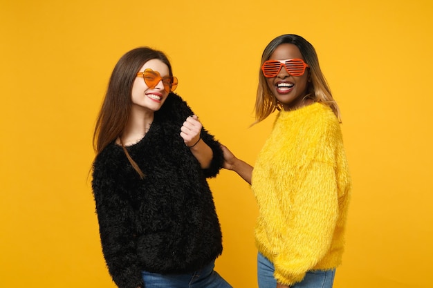 Dois amigos de mulheres jovens europeus e afro-americanos em roupas amarelas pretas em pé posando isolado no fundo da parede laranja brilhante, retrato de estúdio. Conceito de estilo de vida de pessoas. Mock up espaço de cópia.