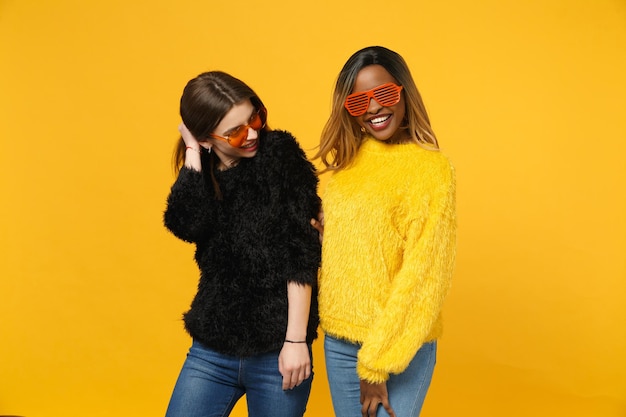 Dois amigos de mulheres jovens europeus e afro-americanos em roupas amarelas pretas em pé posando isolado no fundo da parede laranja brilhante, retrato de estúdio. Conceito de estilo de vida de pessoas. Mock up espaço de cópia.