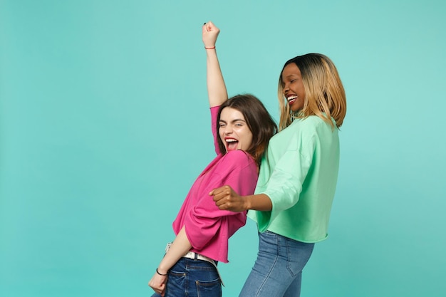 Dois amigos de mulheres jovens europeus e afro-americanos em roupas verdes rosa dançando brincando isolado no fundo da parede azul turquesa, retrato de estúdio. Conceito de estilo de vida de pessoas. Mock up espaço de cópia.