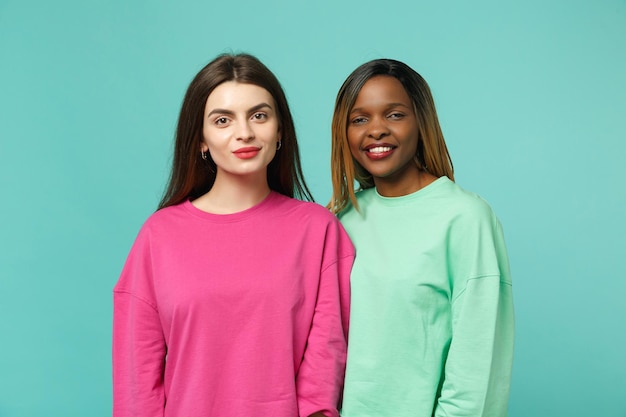 Dois amigos de mulheres jovens europeus e afro-americanos em roupas verdes rosa em pé posando isolado no fundo da parede azul turquesa, retrato de estúdio. Conceito de estilo de vida de pessoas. Mock up espaço de cópia.