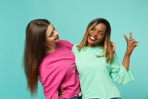 Dois amigos de mulheres jovens europeus e afro-americanos em roupas verdes rosa em pé posando isolado no fundo da parede azul turquesa, retrato de estúdio. Conceito de estilo de vida de pessoas. Mock up espaço de cópia.