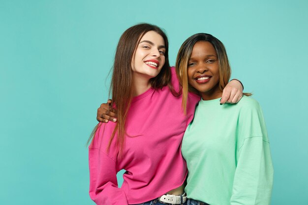 Dois amigos de mulheres jovens europeus e afro-americanos em roupas verdes rosa em pé posando isolado no fundo da parede azul turquesa, retrato de estúdio. Conceito de estilo de vida de pessoas. Mock up espaço de cópia.