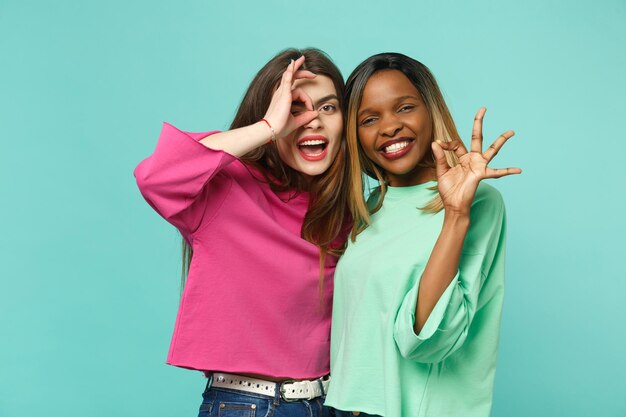 Dois amigos de mulheres jovens europeus e afro-americanos em roupas verdes rosa, mostrando o gesto de OK isolado no fundo da parede azul turquesa, retrato de estúdio. Conceito de estilo de vida de pessoas. Espaço de cópia de mock up