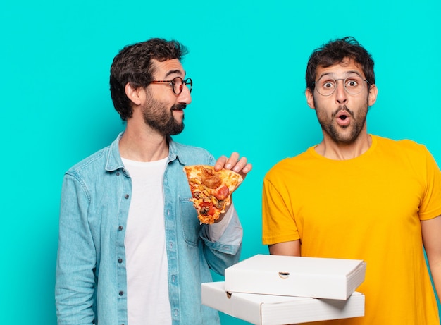 Dois amigos hispânicos com expressão de surpresa e segurando pizzas para viagem