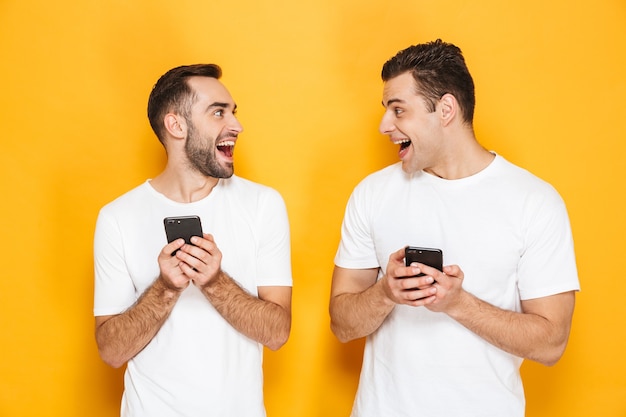 Dois amigos homens alegres e animados, vestindo camisetas em branco, isolados sobre uma parede amarela, usando telefones celulares