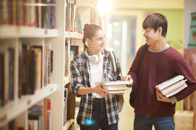 Dois amigos segurando livros sorrindo e conversando enquanto caminham pelas estantes