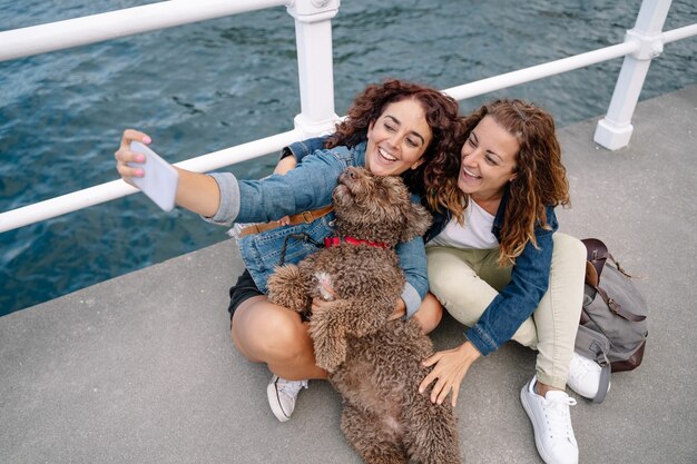 Dois amigos tirando uma foto com o cachorro-d'água marrom. visão horizontal de mulheres viajando com animal de estimação. estilo de vida e tecnologia com animais ao ar livre.