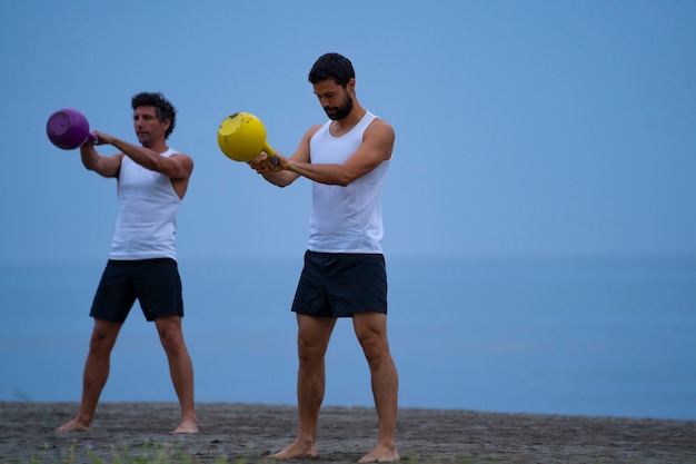 Dois atletas do sexo masculino se exercitando na praia com sinos de chaleira