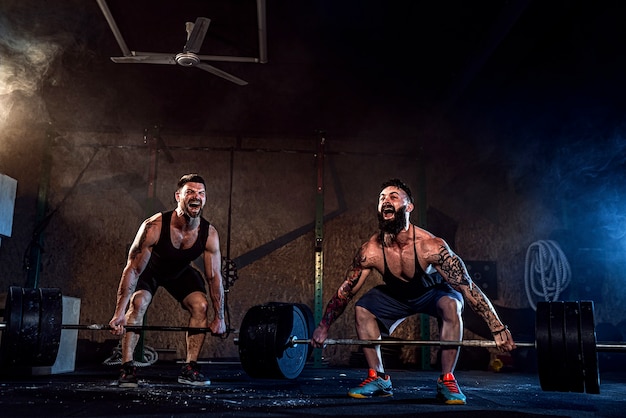 Dois atletas musculosos e tatuados com barbas treinando, levantando pesos pesados na fumaça na academia