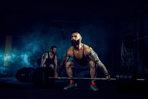 Dois atletas musculosos e tatuados com barbas treinando, um levanta a barra de peso quando o outro está motivando