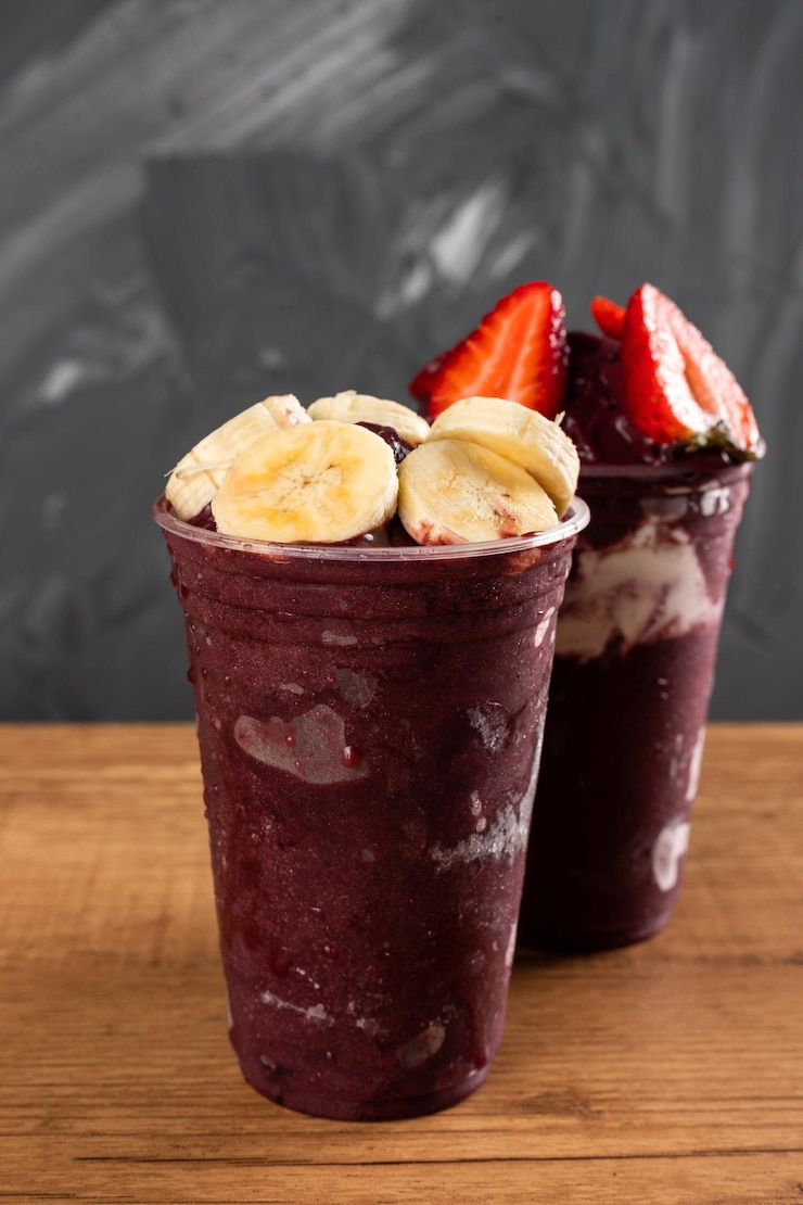 Foto dois batidos de sorvete brasileiro de açaí berry em copo plástico com morangos, bananas e leite condensado. sobre uma mesa de madeira e um fundo cinza de verão. vista frontal para menu e mídia social