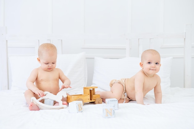 Dois bebês gêmeos meninas de seis meses em fraldas em uma cama de algodão branco em uma roupa de banho em uma cama em casa brincando com brinquedos de madeira e sorrindo