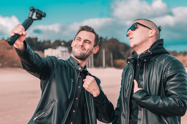Dois bonitos amigos do sexo masculino fazendo Selfie usando a câmera de ação com estabilizador de cardan na praia.
