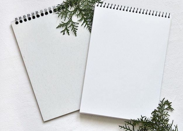 Dois cadernos de desenho brancos sobre uma mola sobre uma toalha de mesa de linho com raminhos de thuja verde