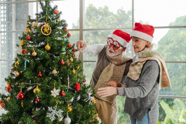 Dois caucasianos sênior casal com camisola e chapéu de papai noel decoram a árvore de natal juntos perto da janela na celebração do festival de natal e ano novo em casa no inverno. atividade de natal e ano novo.