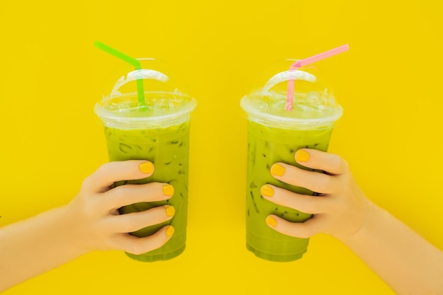 Dois chá verde latte com gelo em copo de plástico e canudo na mão feminina com manicure amarela em fundo amarelo Caseiro Iced Matcha Latte Tea with Milk take away
