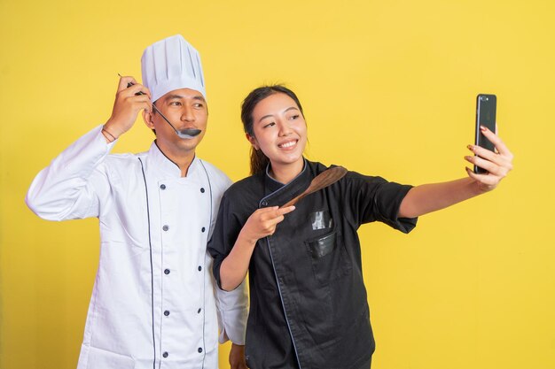 Foto dois chefs segurando utensílios de cozinha enquanto selfie usando telefones celulares em fundo isolado