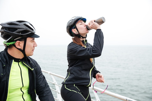 Dois ciclistas descansando perto do mar. mulher bebendo água e homem olhando para o mar