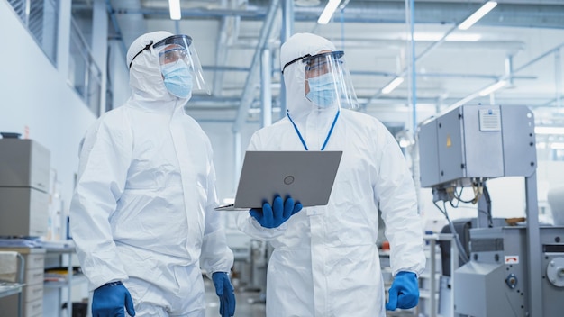 Foto dois cientistas caminhando em uma fábrica de indústria pesada com macacões estéreis e máscaras faciais usando computador portátil examinando configurações de máquinas industriais e configurando a funcionalidade de produção