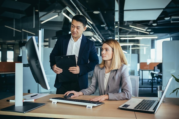 Dois colegas asiáticos masculinos e femininos em um escritório moderno, uma mulher mostra o trabalho realizado no monitor consulta e discute