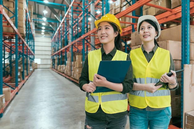 dois colegas de trabalho bem sucedidos jovem usando capacetes e colete de segurança em pé no grande armazém. trabalhadores confiantes de colegas de trabalho enchem de esperanças e energia olhando para cima sorrindo no armazém.