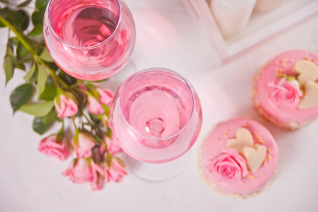 Foto dois copos com vinho de uva rosa com flores rosas e mini bolos. conceito de jantar romântico.