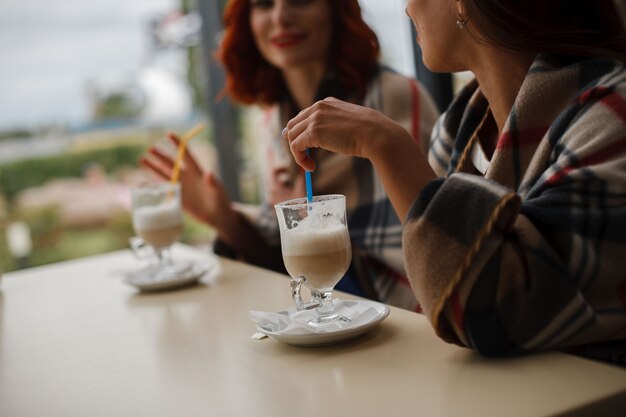 Dois copos de cappuccino em cima da mesa. retrato de duas meninas bebe café no café fechar. copo de cappuccino com um canudo nas mãos da mulher. namoradas falam por uma xícara de café aromático.