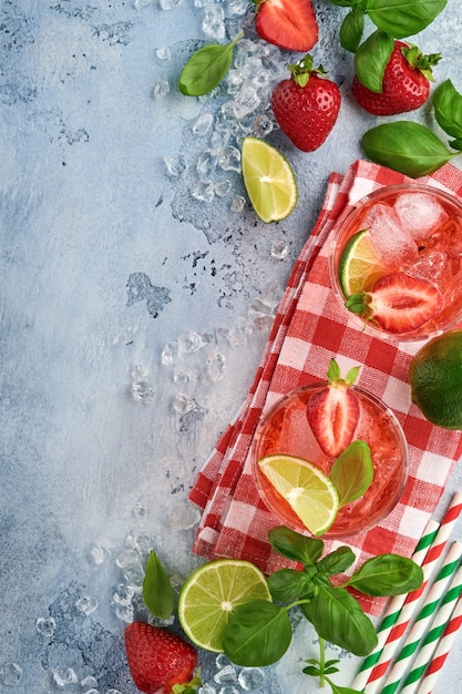 Foto dois copos de ponche e ingredientes frescos para fazer limonada, água desintoxicada com infusão ou coquetel. morangos, limão, hortelã, manjericão, cubos de gelo e agitador em pedra cinza ou fundo de concreto. vista do topo.