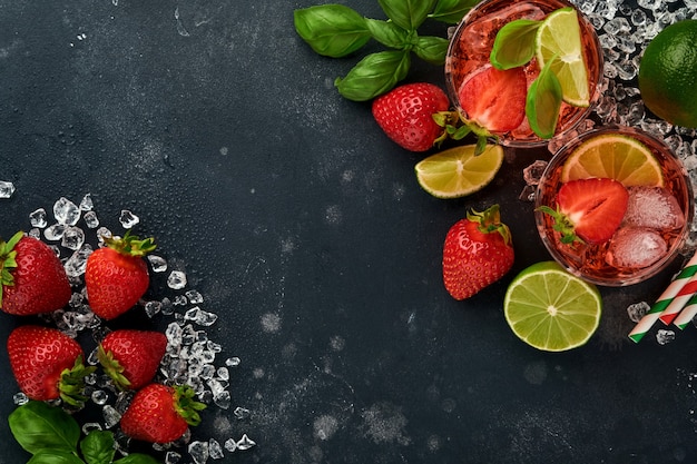 Dois copos de ponche e ingredientes frescos para fazer limonada, água desintoxicante com infusão ou coquetel. Morangos, limão, hortelã, manjericão, cubos de gelo e agitador em pedra preta ou fundo de concreto. Vista do topo.