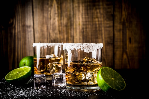 Dois copos de tequila na mesa escura, com cubos de gelo, sal e limão