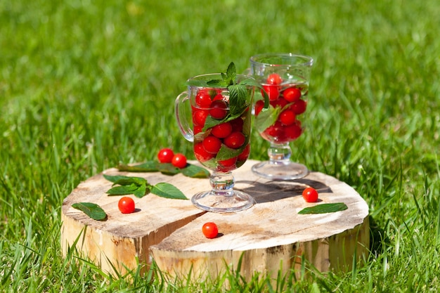 Dois copos de vidro com frutas cereja, hortelã e água ficam em um carrinho de madeira com uma rachadura no gramado verde em um dia ensolarado de verão.