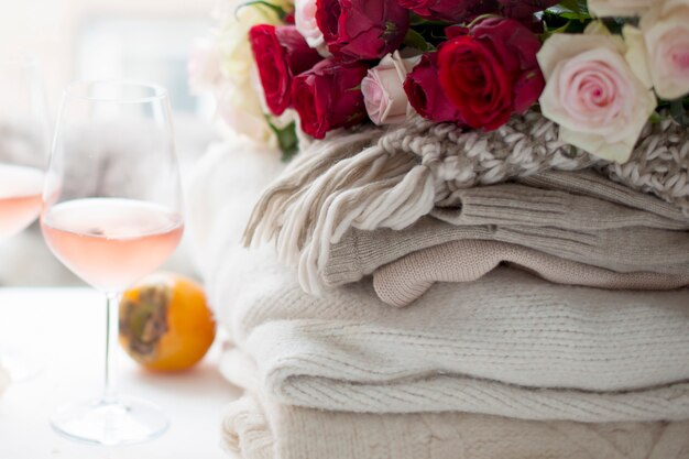 Dois copos de vinho e um monte de roupas quentes de inverno em casa perto da janela. gato, grande buquê de rosas e frutas de outono no quadro. a cidade está fora da janela. espaço livre para texto.