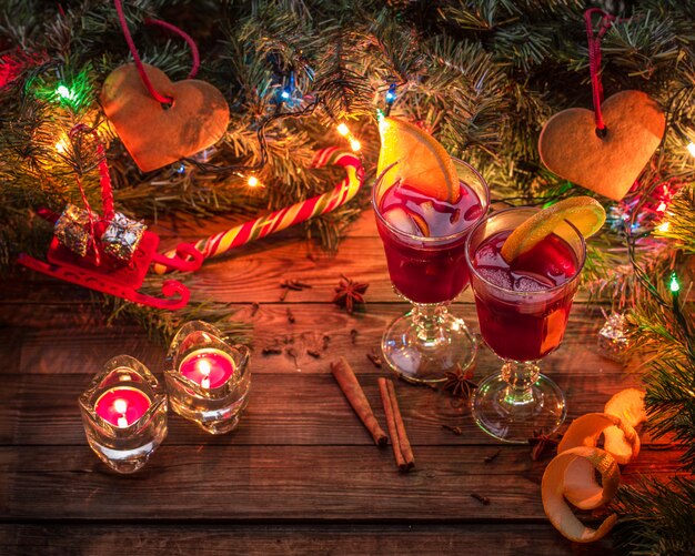 Dois copos de vinho quente com fatias de laranja, velas e árvore de Natal com guirlanda e brinquedos