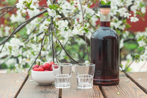 Dois copos vazios e uma garrafa de licor de cereja sobre uma árvore em flor. Álcool caseiro feito de frutas vermelhas.