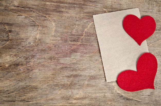 Dois corações de tecido vermelho com folha de papel deitada na velha mesa de madeira.