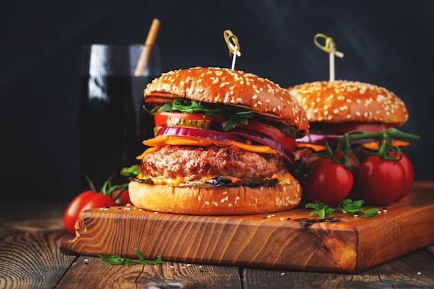 Dois deliciosos hambúrgueres caseiros de carne, queijo e legumes em uma velha mesa de madeira. Close de alimentos gordurosos e pouco saudáveis.