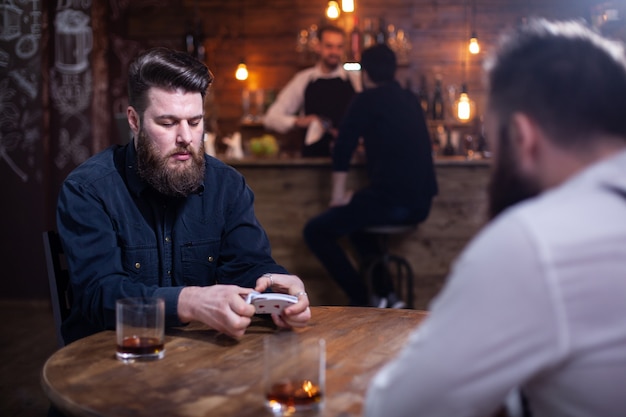 Dois descolados barbudos jogando cartas em um pub vintage. Um barman com um cliente está em segundo plano