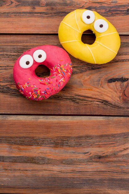 Dois donuts surpresos sobre fundo de madeira marrom. Rosquinhas rosa e amarelas com olhos engraçados. Vista superior, copie o espaço.