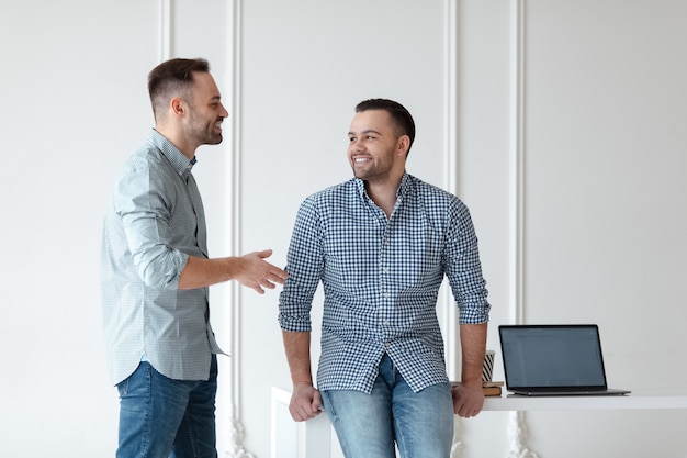 Dois empresários bem sucedidos e sorridentes, falando sobre trabalho, na superfície da parede branca. Laptop, café e notebook na mesa de escritório branca.