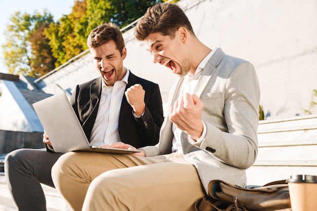 Dois empresários confiantes usando um laptop enquanto estão sentados em um banco ao ar livre, comemorando o sucesso