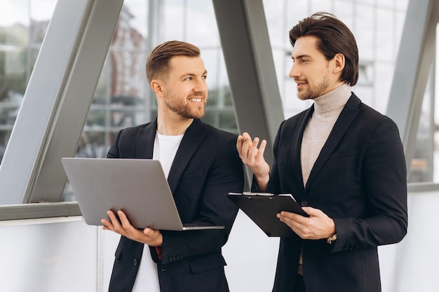 Dois empresários felizes modernos com laptop e pasta de documentos discutindo algo no contexto de escritórios e edifícios urbanos