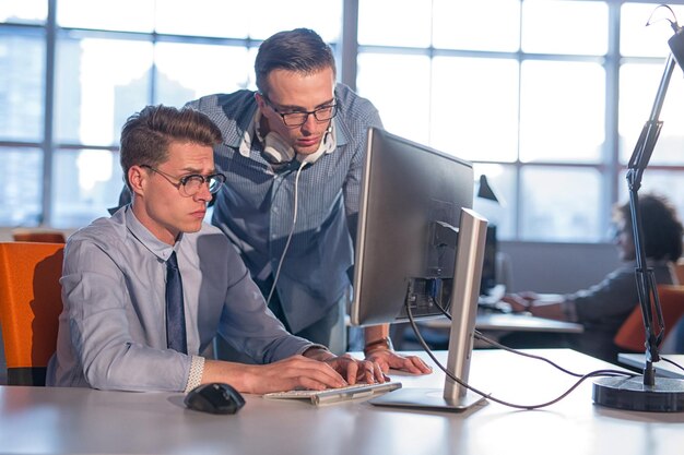 Dois empresários usando computador se preparando para a próxima reunião e discutindo ideias com colegas em segundo plano