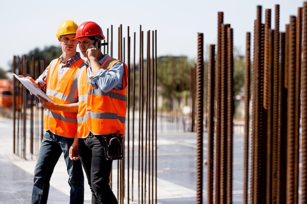 Dois engenheiros estruturais vestidos com camisas, coletes de trabalho laranja e capacetes exploram a documentação da construção e conversam por telefone no canteiro de obras próximo às estruturas de aço.