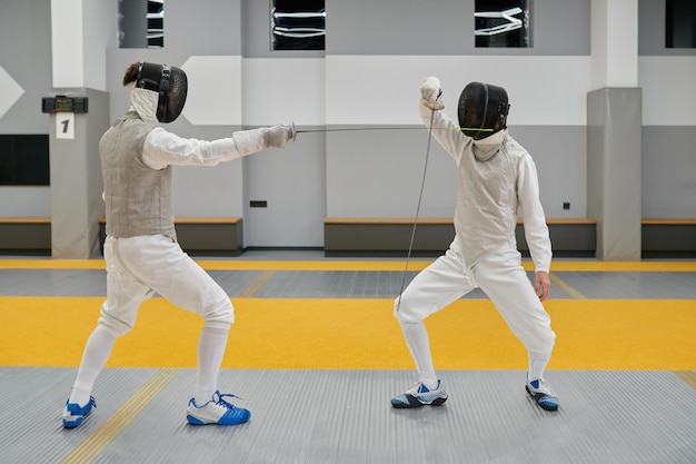 Dois espadachins concentrados vestindo uniforme e máscara protetora exercitando e lutando com floretes durante a competição de esgrima