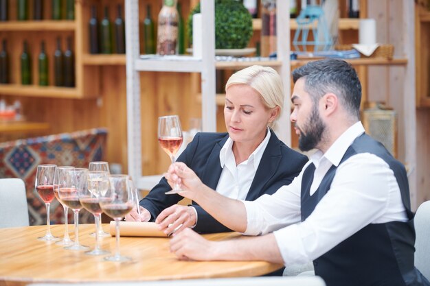 Dois especialistas em vinícolas discutindo características de novos tipos de vinho na adega enquanto um deles faz anotações