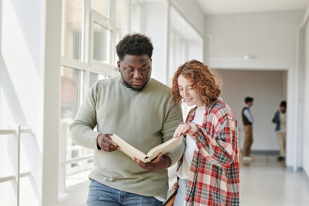 Dois estudantes adolescentes interraciais lendo livro da biblioteca no corredor da faculdade