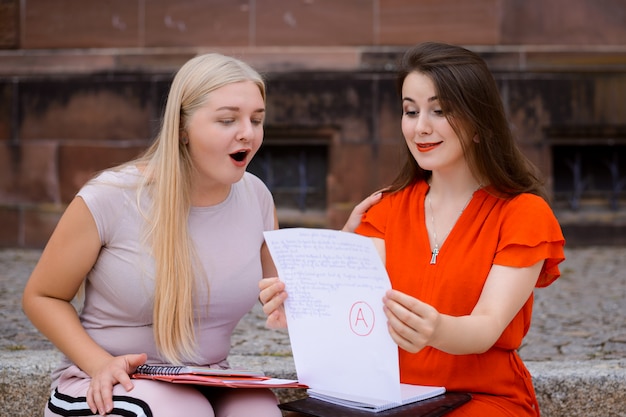 Dois estudantes animados olhando para papel de exame juntos sentados perto da universidade