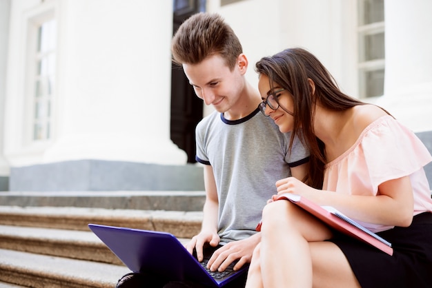 Dois estudantes felizes usando laptop moderno trabalhando no projeto, preparam-se para as aulas nas escadas da universidade convencional antes das aulas