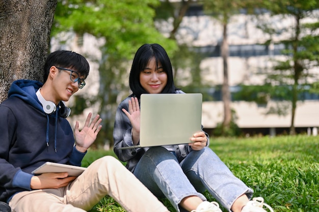 Dois estudantes universitários asiáticos sorridentes estão tendo uma reunião online enquanto estão sentados no parque